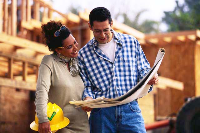 A homeowner and a contractor read building plans together outside a house being framed. A yellow hard hat with PAC's green check sits in the foreground.
